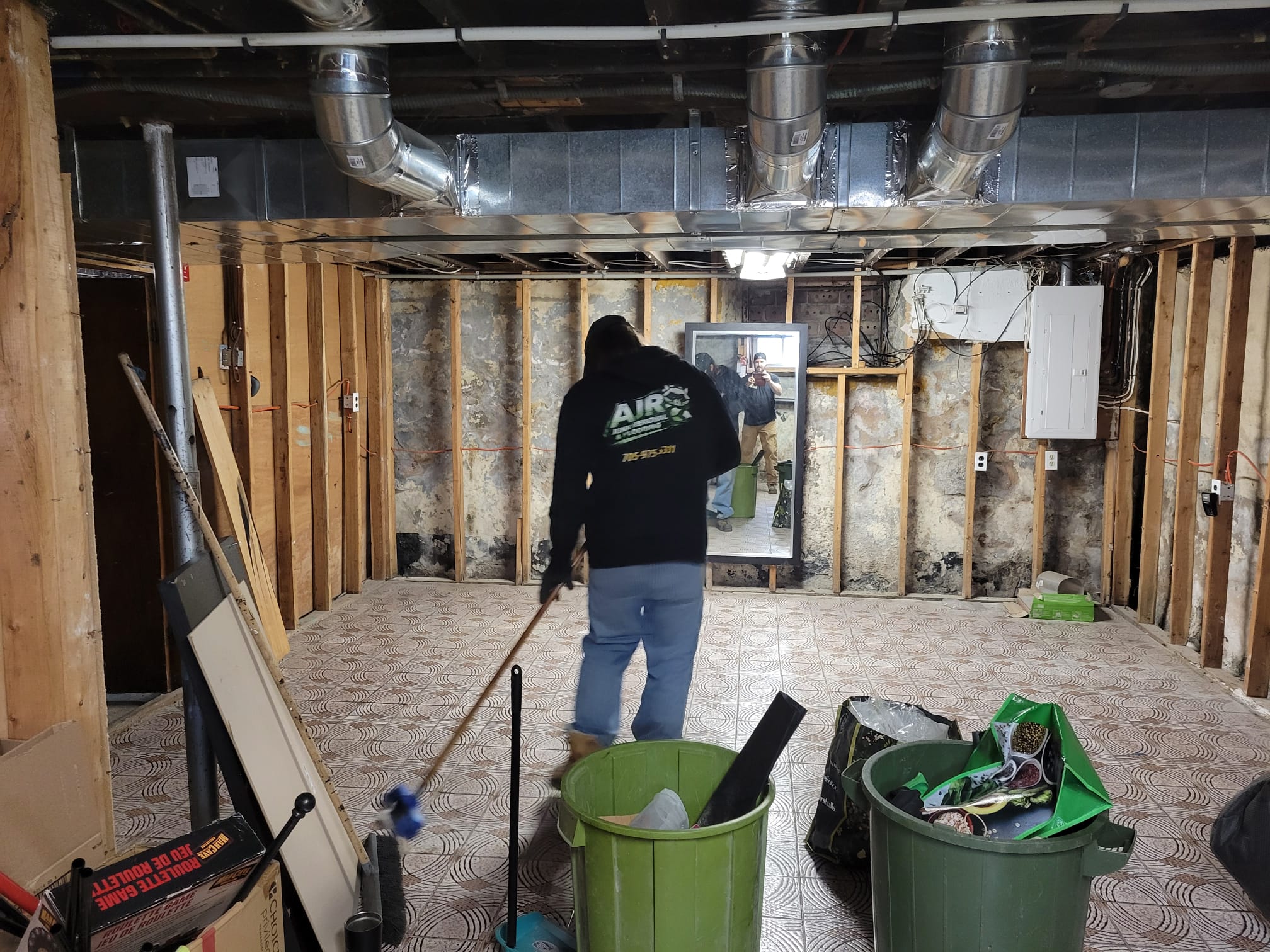 A worker wearing a black AJR Junk Removal & Flooring hoodie sweeping a tiled floor in a gutted basement with exposed wooden wall studs and ductwork.