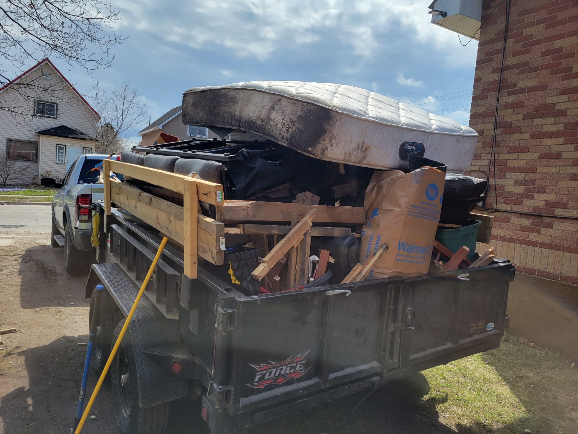 A black dump trailer parked in a residential driveway, completely filled with debris including a stained mattress, a black leather couch, broken wood, and yard waste.