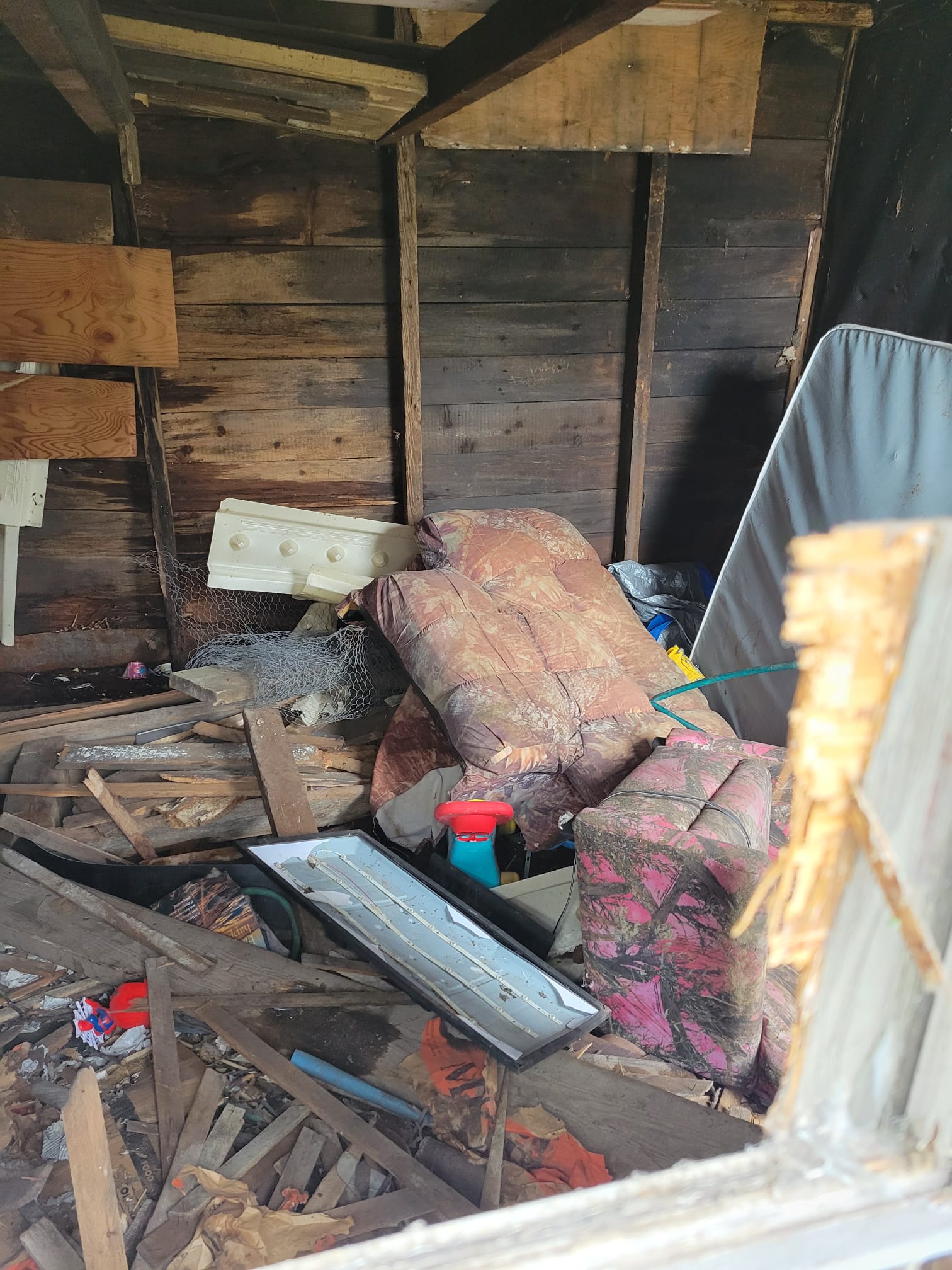 The interior of a dilapidated wooden shed filled with piled-up junk, including old mattresses, broken wood planks, a pink camo-patterned cushion, a shattered light fixture, and rolled-up chicken wire.